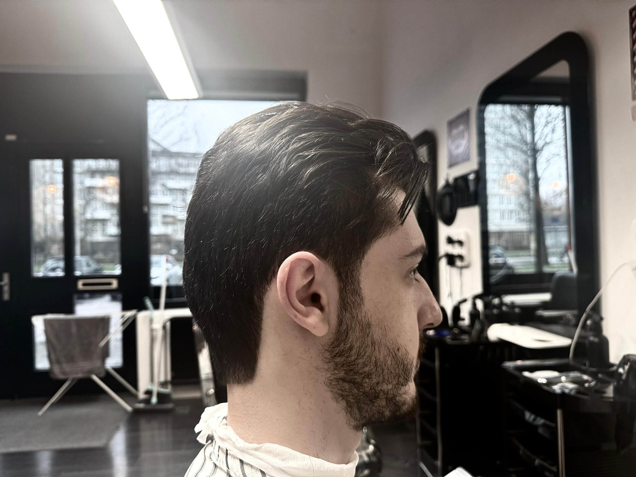 Klant met getaperd kapsel en baard van opzij, met de spiegel en de ramen van de zaak achter hem